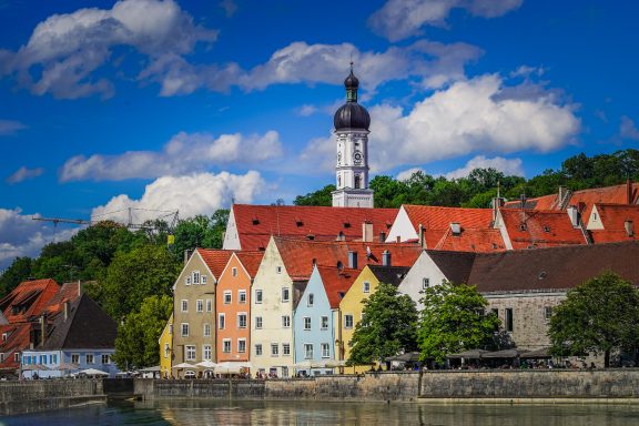 Bunte Häuser mit roten Dächern und Kirchturm am Wasser, umgeben von Bäumen und blauem Himmel.