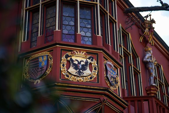 Fassade eines Gebäudes mit Wappen und dekorativen Elementen in Rottönen.